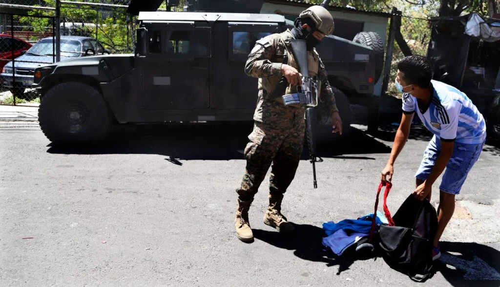 El Salvadorian soldier holding an assault rifle checks another man's bag on the ground